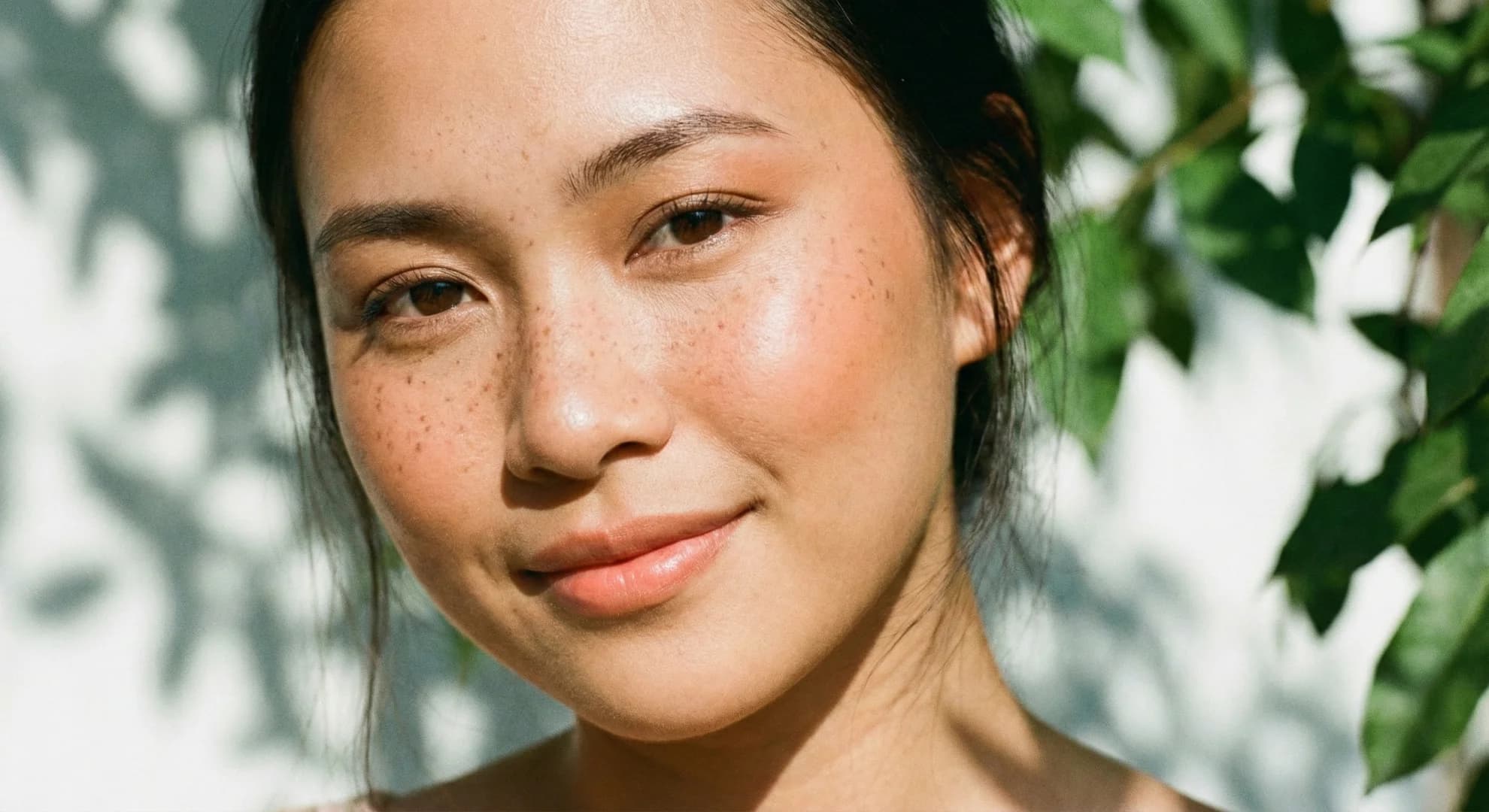 Close-up of a smiling woman with freckles and glowing skin in natural sunlight.