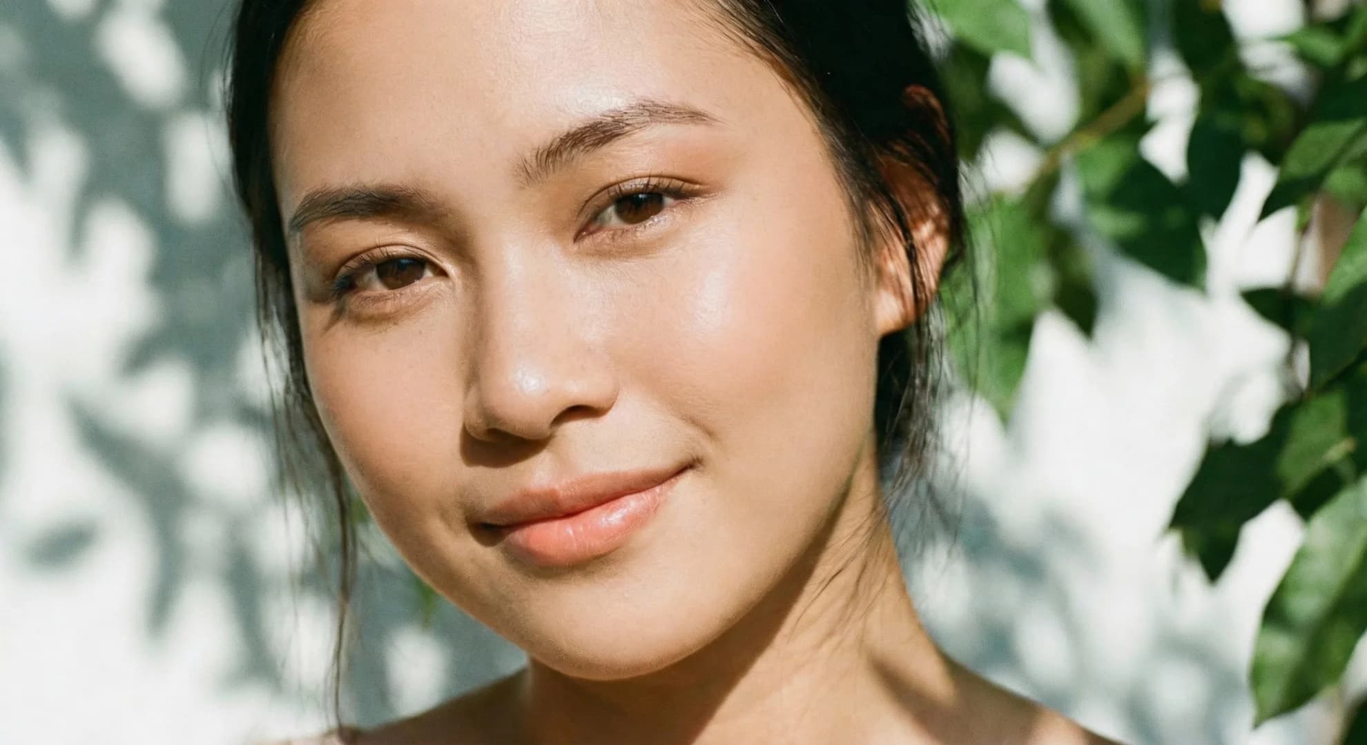 Close-up of a smiling woman with glowing skin, framed by soft sunlight and green leaves.